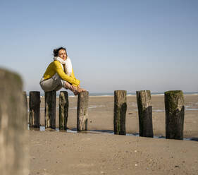Woman sitting on wooden post at beach against clear sky during sunny day - UUF22334