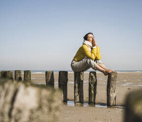 Mature woman sitting on wooden post at beach against clear sky during sunny day - UUF22335