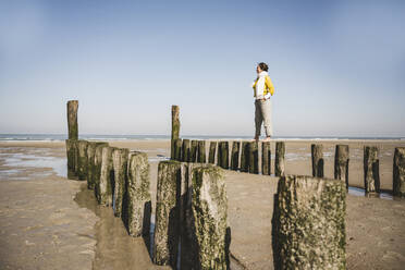 Woman standing on wooden post at beach against clear sky during sunny day - UUF22338