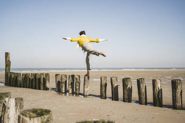Woman with arms outstretched balancing on wooden posts at beach against clear sky - UUF22339