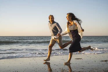 Fröhliches junges Paar, das sich beim Laufen am Strand während des Sonnenuntergangs an den Händen hält - UUF22357