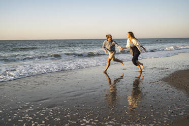 Glückliches Paar hält Hände beim Laufen am Strand gegen den klaren Himmel bei Sonnenuntergang - UUF22358