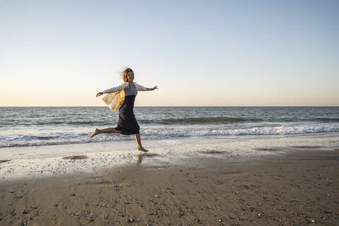 Cheerful young woman running at beach against clear sky during sunset stock photo