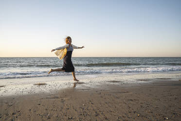 Cheerful young woman running at beach against clear sky during sunset - UUF22361