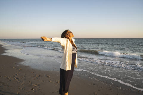 Carefree young woman standing at beach with arms outstretched during sunset stock photo