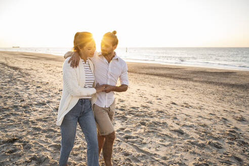 Junges Paar hält Hände beim Spaziergang am Strand während eines sonnigen Tages - UUF22367