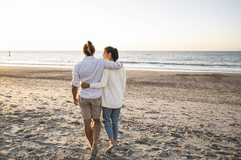 Junges Paar, das sich in den Armen liegt und bei Sonnenuntergang am Strand spazieren geht - UUF22369