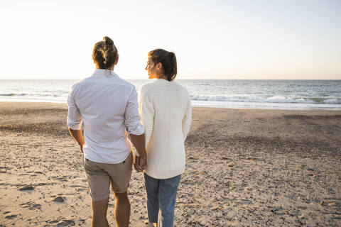 Young couple holding hands while walking at beach during sunset stock photo