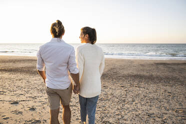 Young couple holding hands while walking at beach during sunset - UUF22370
