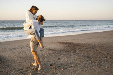 Carefree young man lifting girlfriend at beach during sunset - UUF22374