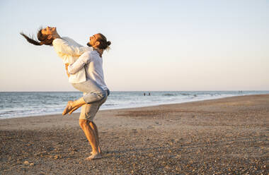 Carefree boyfriend lifting girlfriend at beach against clear sky during sunset - UUF22375