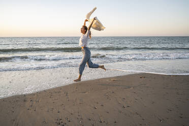 Carefree woman running at beach against clear sky during sunset - UUF22377