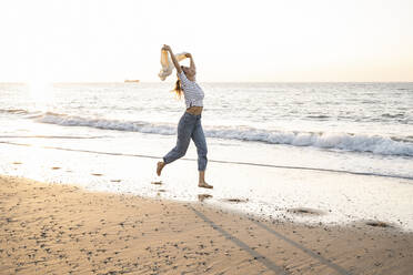 Carefree young woman holding jacket while running at beach against clear sky during sunset - UUF22378