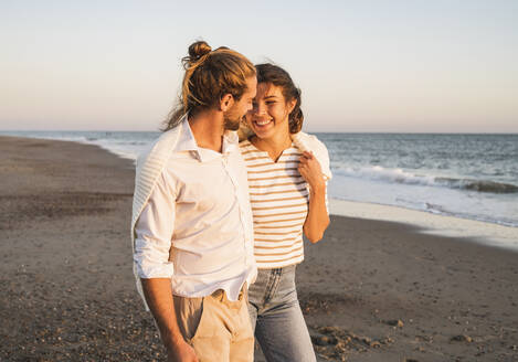 Lächelnde junge Frau, die mit ihrem Freund am Strand bei Sonnenuntergang spazieren geht - UUF22380
