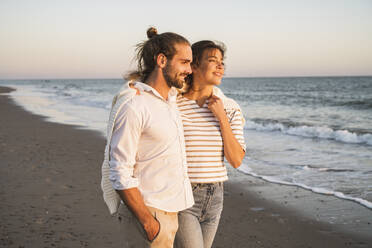 Lächelndes junges Paar schaut weg, während es im Urlaub am Strand spazieren geht - UUF22381
