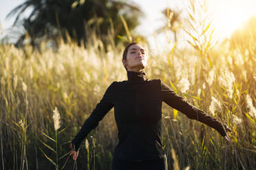 Young woman with eyes closed standing against plants during sunset - MPPF01352