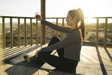 Junge Frau, die ein Selfie mit ihrem Smartphone macht, während sie bei Sonnenuntergang auf einer Matte sitzt - MPPF01356