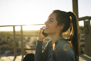 Lächelnde Frau, die über ihr Smartphone spricht, während sie gegen den klaren Himmel bei Sonnenuntergang sitzt - MPPF01357