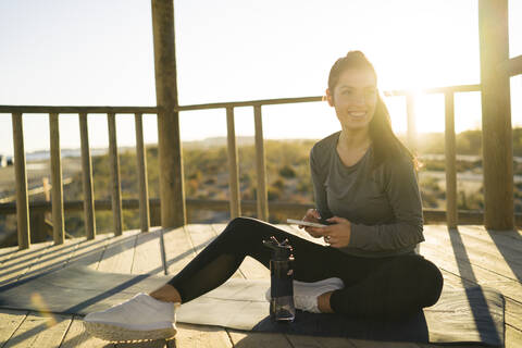 Lächelnde junge Frau, die ein Smartphone benutzt, während sie auf einer Matte im Pavillon sitzt, lizenzfreies Stockfoto