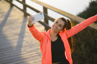 Lächelnde junge Frau, die ein Selfie mit ihrem Mobiltelefon macht, während sie auf einer Fußgängerbrücke steht - MPPF01364