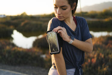 Junge Frau, die ihr Smartphone am Armring einstellt, während sie vor einer Landschaft steht - MPPF01365