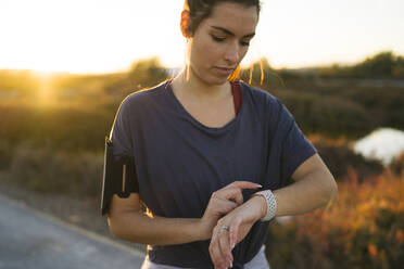 Junge Frau benutzt eine intelligente Uhr, während sie bei Sonnenuntergang vor der Landschaft steht - MPPF01366