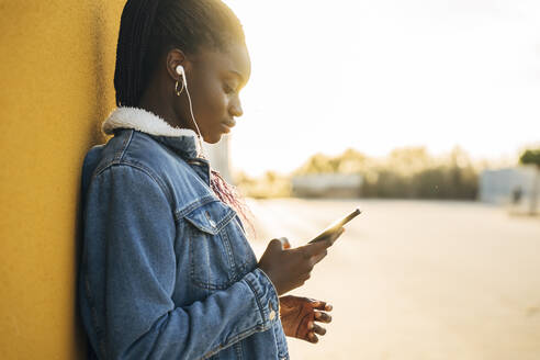 Teenager-Mädchen mit Smartphone beim Hören von Musik durch gelbe Wand auf der Straße - MPPF01384