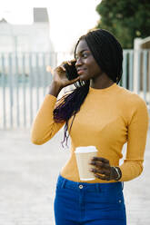 Smiling teenage girl with coffee cup looking away while talking on smart phone at street - MPPF01405
