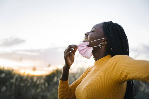 Happy teenage girl with eyes closed removing mask and inhaling in field during sunset - MPPF01417