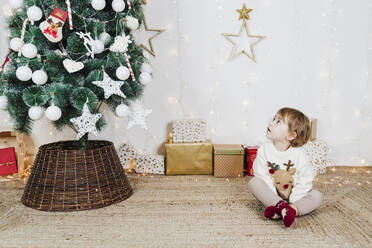 Ein kleines Mädchen betrachtet den Weihnachtsbaum, während es auf einem Teppich an der Wand sitzt - EBBF01953
