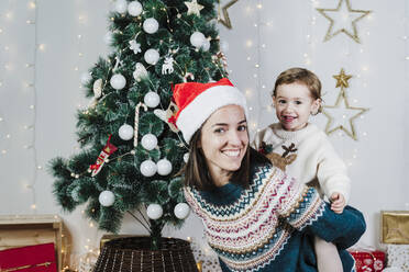 Lächelnde Mutter mit Weihnachtsmannmütze, die ihre süße Tochter zu Hause während der Weihnachtszeit huckepack nimmt - EBBF01964