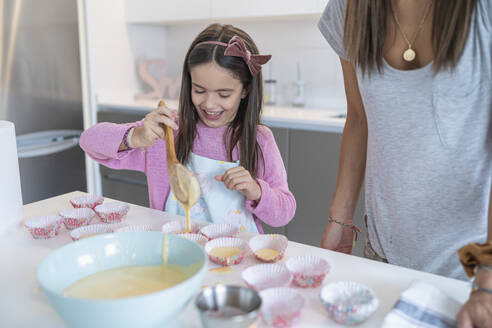 Glückliche Tochter schöpft Teig in Muffinhalter von Mutter in Küche - SNF00893