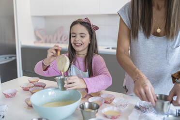 Lächelnde Tochter schöpft Teig in Cupcake-Halter von Mutter in Küche - SNF00896