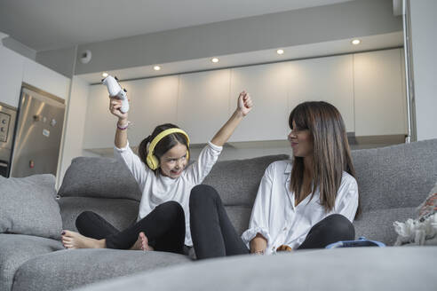 Cheerful daughter with hand raised sitting by mother while playing video games in living room - SNF00921