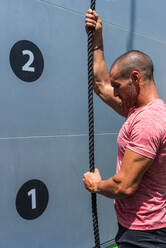 Side view of confident male athlete climbing rope while doing exercises on sports ground in summer on sunny day - ADSF19662