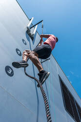 From below of confident male athlete climbing rope while doing exercises on sports ground in summer on sunny day - ADSF19663