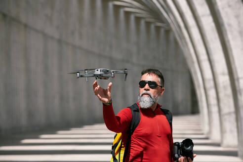 Elderly male photographer standing in archway on sunny day with modern flying drone in La Palma - ADSF19677