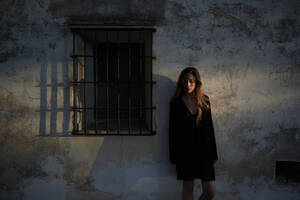 Ganzkörperdarstellung einer jungen Frau in legerem schwarzen Kleid und Stiefeln, die sich an die schäbige Steinmauer eines alten Gebäudes mit kleinem Fenster lehnt - ADSF19714