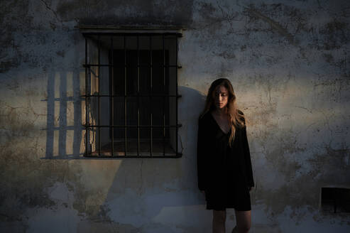 Full body of young female in casual black dress and boots leaning back against shabby stone wall of aged building with small window - ADSF19714