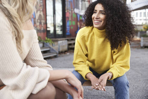 Smiling young woman talking with female friend while sitting on seat - SUF00658