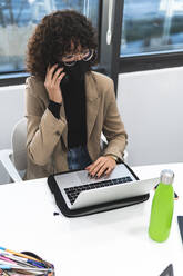 Businesswoman working on laptop while taking phone call in office during COVID-19 - JAQF00052
