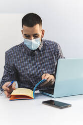 Young businessman with laptop checking diary on desk against white wall during COVID-19 - JAQF00055