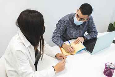 Businessman writing in diary while working with female coworker on desk during COVID-19 - JAQF00060