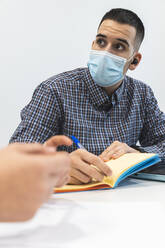 Businessman wearing protective face mask writing in diary during meeting with colleague at office - JAQF00078