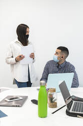 Male entrepreneur looking at female colleague while working against wall at work place during COVID-19 - JAQF00081