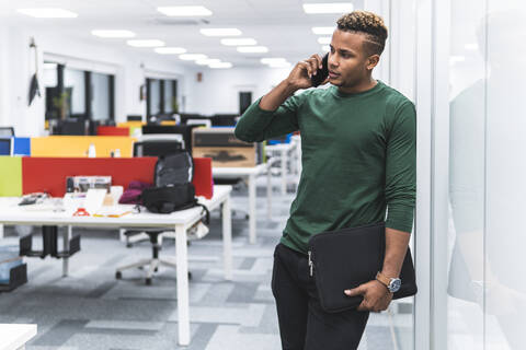 Männlicher Berufstätiger mit Aktenordner bei einem Telefongespräch, der sich bei der Arbeit an ein Glas lehnt, lizenzfreies Stockfoto