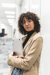 Selbstbewusste weibliche Fachkraft mit Laptop an der Glaswand lehnend am Arbeitsplatz - JAQF00093
