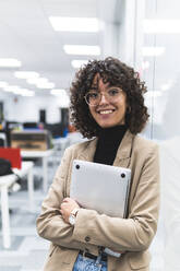 Glückliche Geschäftsfrau mit Laptop vor einer Glaswand am Arbeitsplatz - JAQF00095