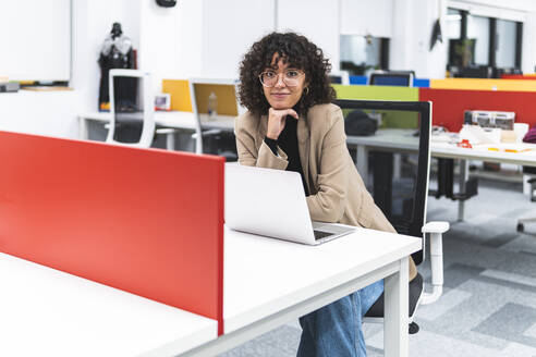 Lächelnde weibliche Fachkraft mit Hand am Kinn am Schreibtisch am Arbeitsplatz - JAQF00099