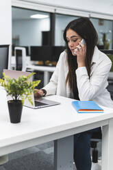 Unternehmerin arbeitet am Laptop, während sie einen Anruf am Schreibtisch im Büro entgegennimmt - JAQF00107